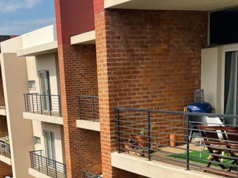 A private balcony featuring a metal balustrade and sliding glass door access, set against an exposed brick exterior facade of the apartment building.