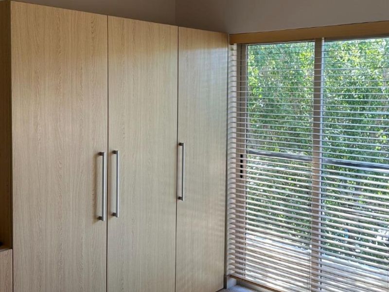A bedroom featuring light wood-grain built-in wardrobes and venetian blinds over a window with a leafy outlook.