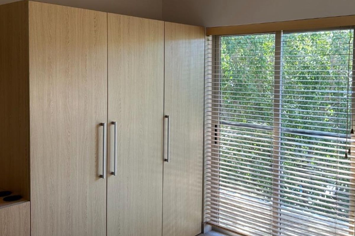 A bedroom featuring light wood-grain built-in wardrobes and venetian blinds over a window with a leafy outlook.