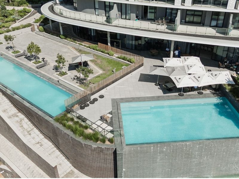 Aerial view of a modern apartment complex featuring tiered swimming pools, paved outdoor entertainment areas, and landscaped gardens with glass balustrades.