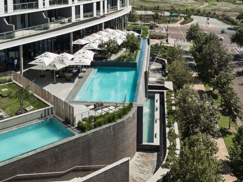 Elevated view of a modern apartment complex featuring tiered swimming pools, landscaped garden areas, and paved outdoor terrace spaces.
