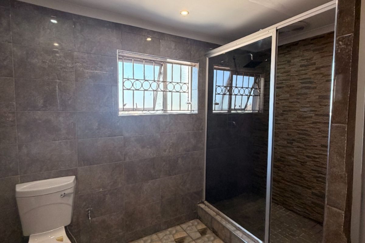A functional bathroom featuring a walk-in shower, tiled finishes, and a toilet, with recessed lighting and security bars on the window.
