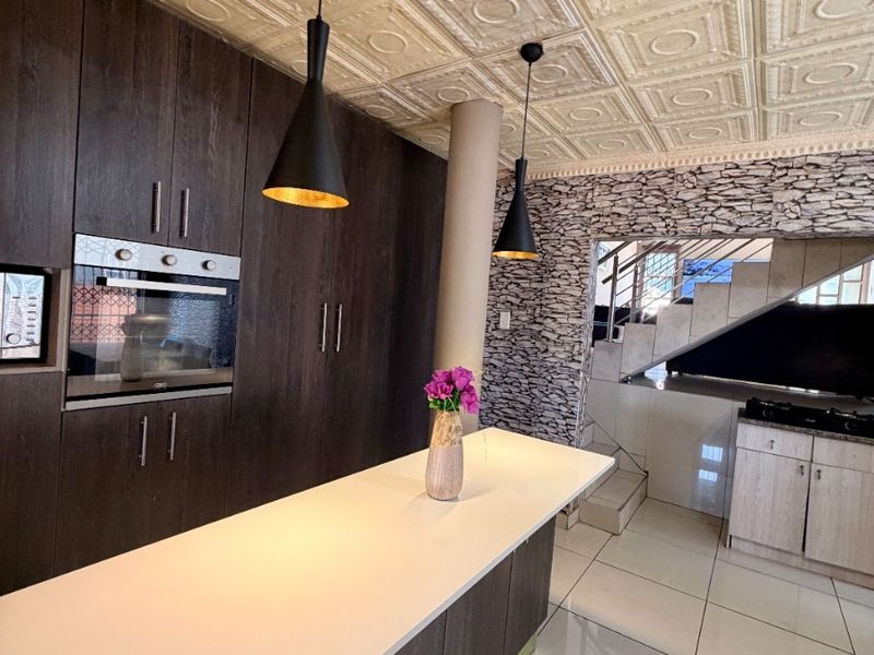 Modern kitchen featuring dark cabinetry, a central island, stone-cladding accent wall, and unique pressed metal ceiling detail, adjacent to a staircase.