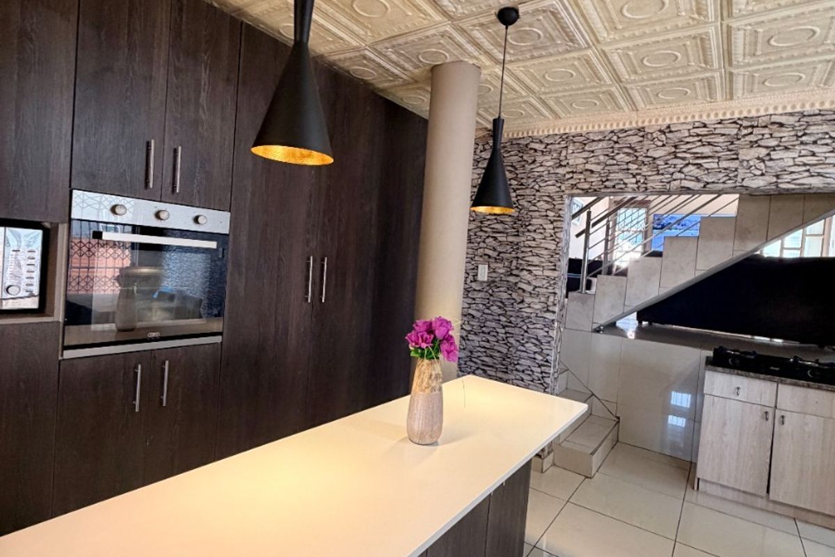 Modern kitchen featuring dark cabinetry, a central island, stone-cladding accent wall, and unique pressed metal ceiling detail, adjacent to a staircase.