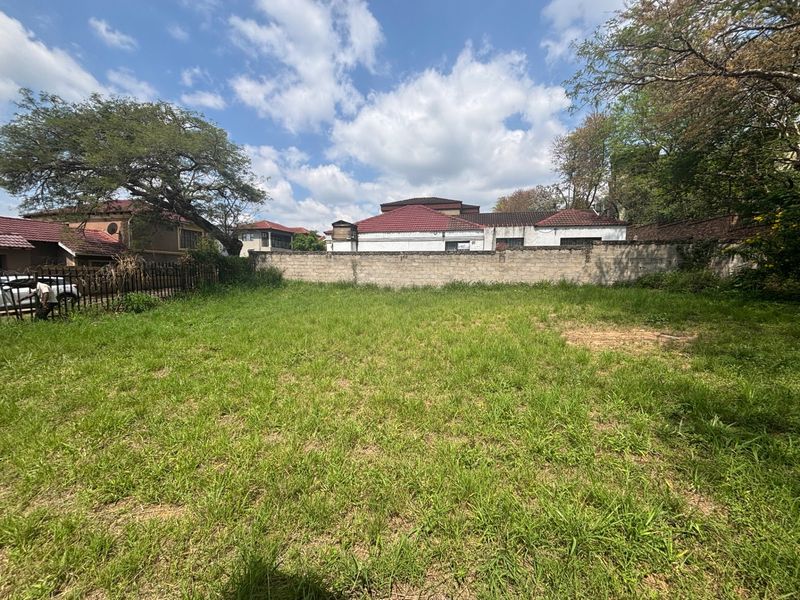 A vacant residential plot featuring a perimeter masonry wall and metal fencing, currently covered in overgrown grass.