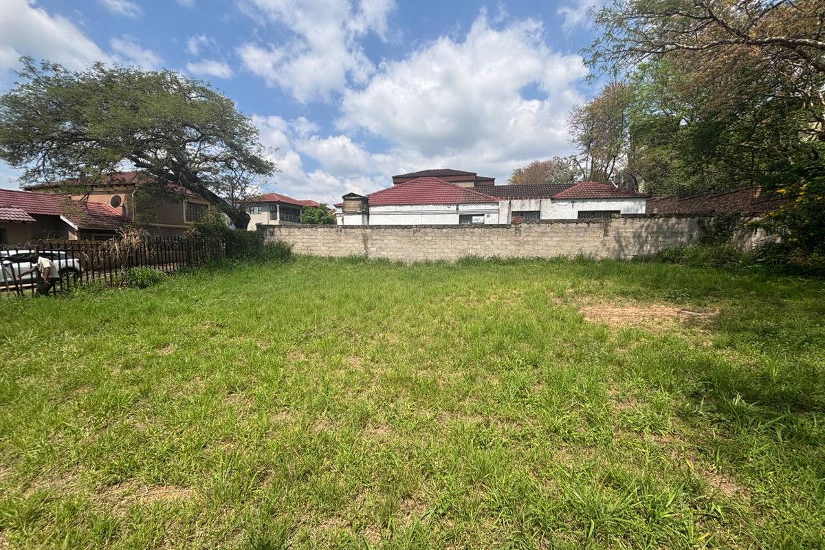 A vacant residential plot featuring a perimeter masonry wall and metal fencing, currently covered in overgrown grass.