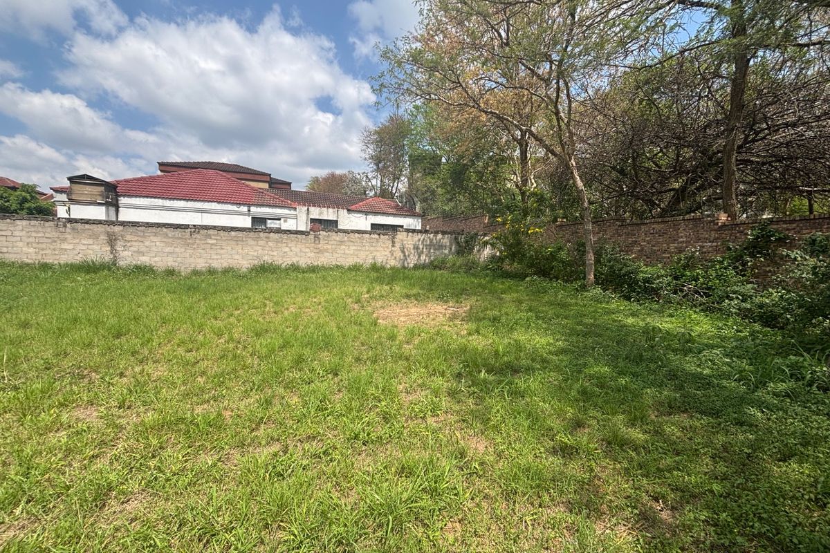 A vacant residential plot featuring a grassed area enclosed by a perimeter brick wall and mature trees.