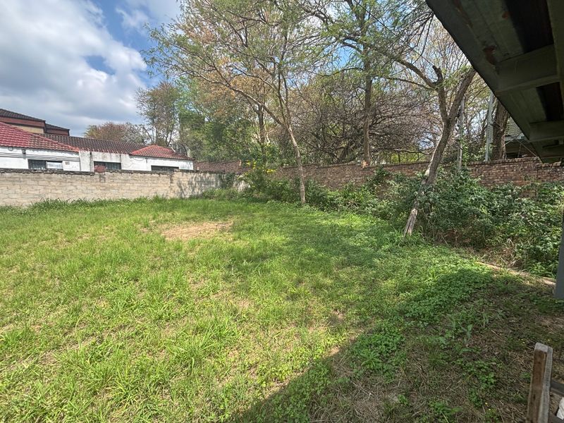A spacious, grassed backyard area enclosed by a brick boundary wall with mature trees, offering potential for landscaping or development.