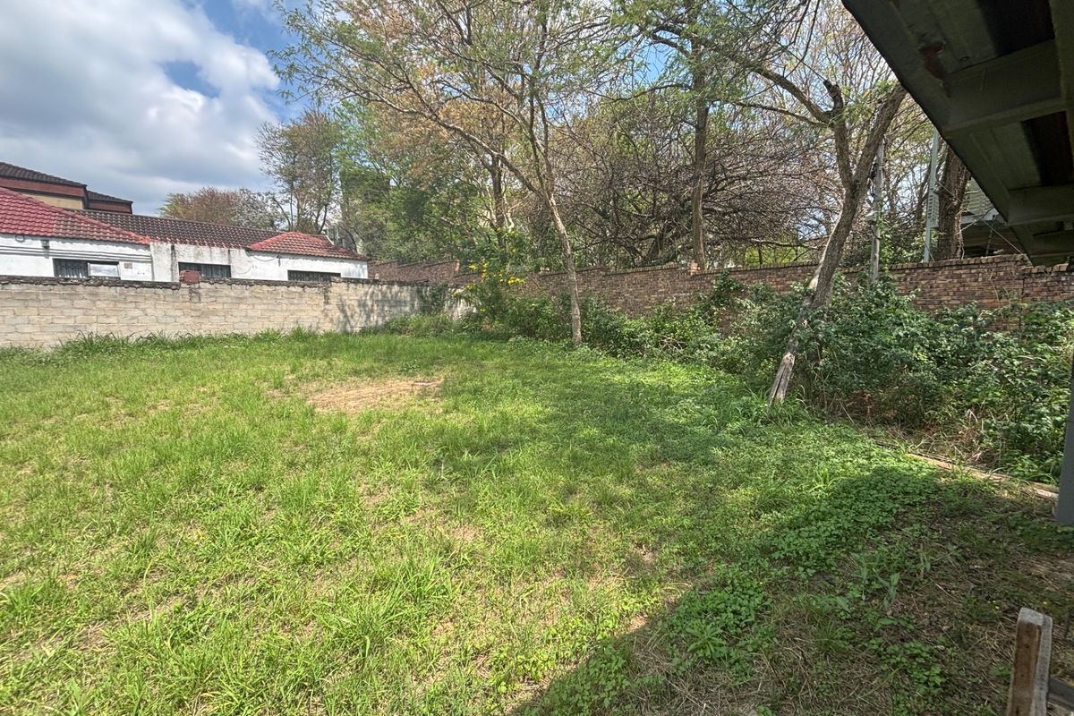 A spacious, grassed backyard area enclosed by a brick boundary wall with mature trees, offering potential for landscaping or development.