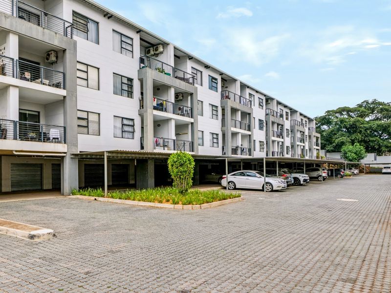 Exterior view of a multi-story apartment complex featuring covered parking bays, paved driveway, and private balconies for each unit.
