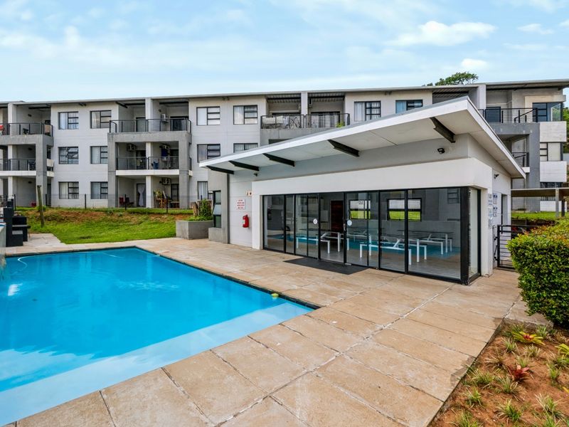 Communal swimming pool area featuring a paved deck and a modern clubhouse building with glass frontage, set against an apartment complex.