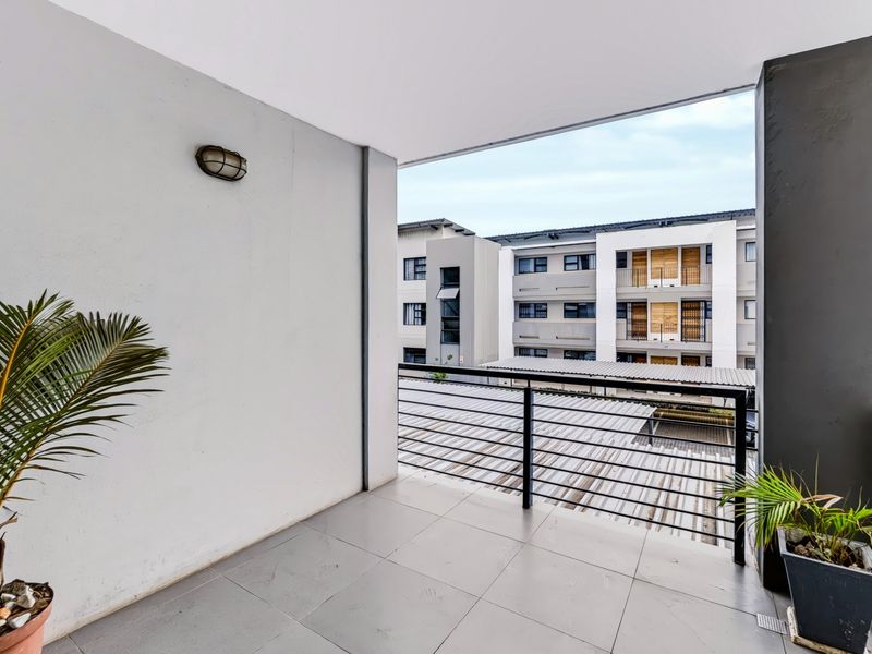 A clean, covered balcony featuring tiled flooring and a metal balustrade, offering an open view of the surrounding apartment complex.