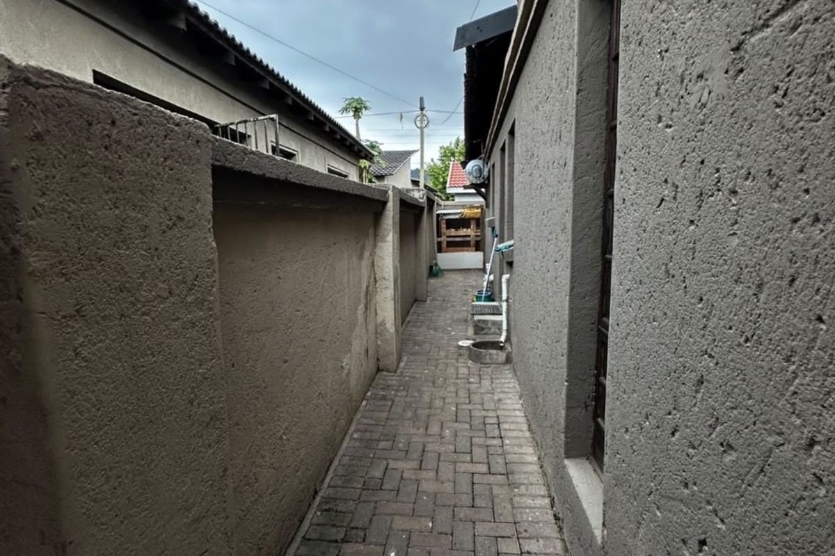 A narrow side passage featuring paved brick flooring and high boundary walls, providing access between the main structure and the rear of the property.