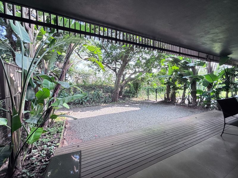 A covered patio featuring timber decking and a slatted ceiling screen, overlooking a private gravel garden area with mature trees.