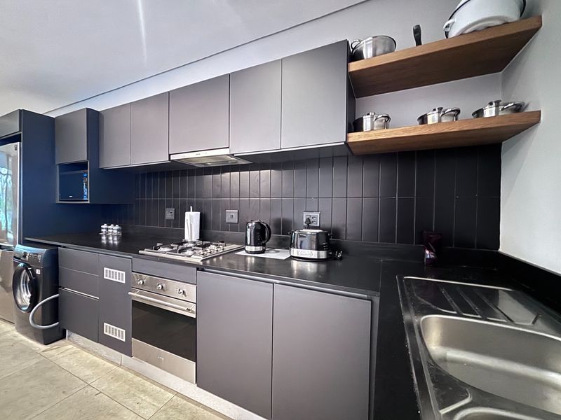 Modern kitchen featuring matte grey cabinetry, black tiled backsplash, gas hob, built-in oven, and integrated laundry appliance with sleek wooden shelving.