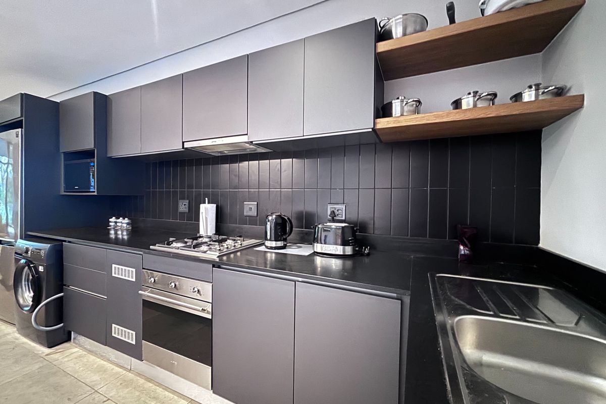 Modern kitchen featuring matte grey cabinetry, black tiled backsplash, gas hob, built-in oven, and integrated laundry appliance with sleek wooden shelving.