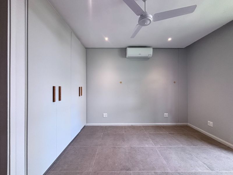 A clean, empty bedroom featuring neutral tiled flooring, built-in cupboards, a ceiling fan, and a wall-mounted air conditioning unit.