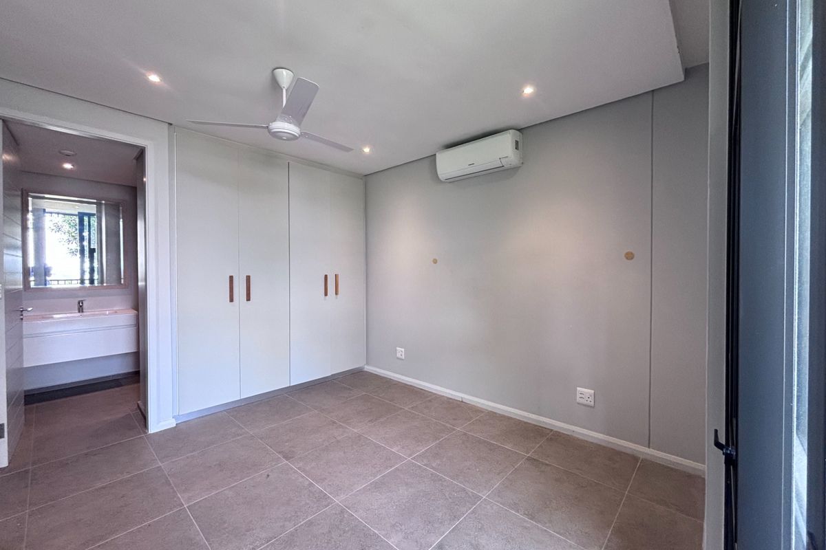 A clean, modern bedroom featuring neutral tiled flooring, built-in cupboards, a ceiling fan, and a wall-mounted air conditioning unit with an adjoining en-suite bathroom.