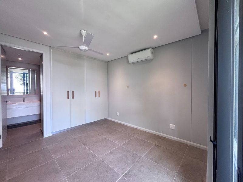 A clean, modern bedroom featuring neutral tiled flooring, built-in cupboards, a ceiling fan, and a wall-mounted air conditioning unit with an adjoining en-suite bathroom.