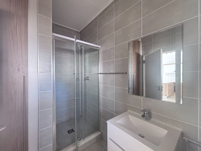 A modern, clean bathroom featuring a glass-enclosed shower, a vanity basin with a wall-mounted mirror, and neutral grey wall tiling.