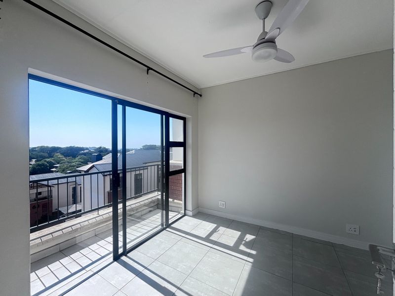 A bright, empty bedroom featuring tiled flooring, a ceiling fan, and a large sliding glass door leading to a private balcony.