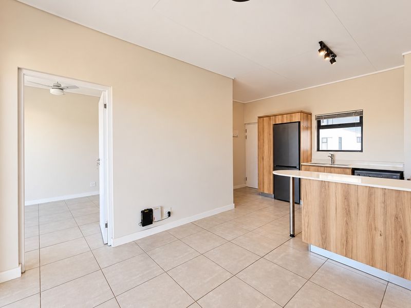 Open-plan living and kitchen area featuring modern wood-finish cabinetry, a breakfast bar, tiled flooring, and an adjoining room with a ceiling fan.