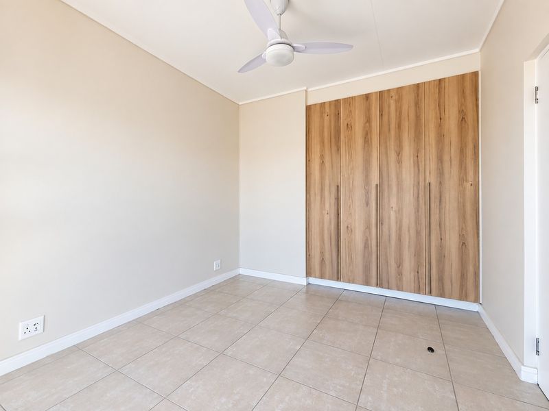 A bright, empty bedroom featuring modern wood-finish built-in cupboards, neutral tiled flooring, and a ceiling fan.