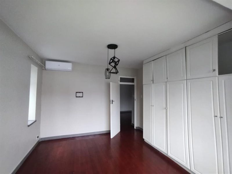 A bright, empty bedroom featuring dark laminate flooring, ample built-in cupboard storage, and a wall-mounted air conditioning unit.