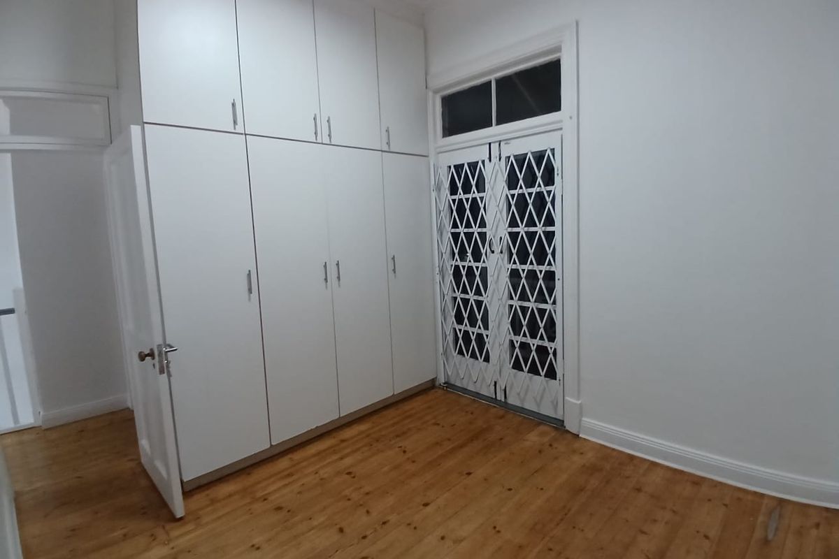 A bright, empty bedroom featuring polished timber flooring, extensive floor-to-ceiling built-in cupboards, and a secure double-door entrance with a fitted security gate.