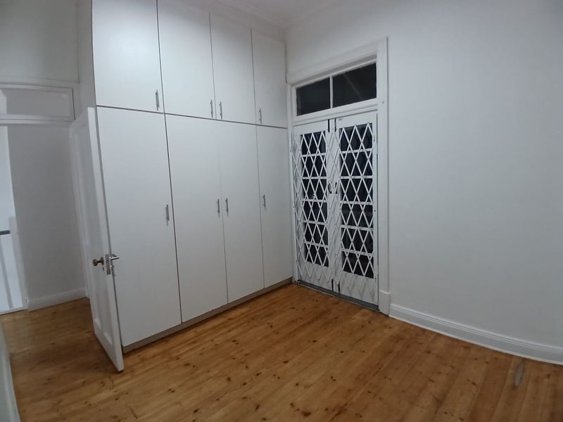 A bright, empty bedroom featuring polished timber flooring, extensive floor-to-ceiling built-in cupboards, and a secure double-door entrance with a fitted security gate.