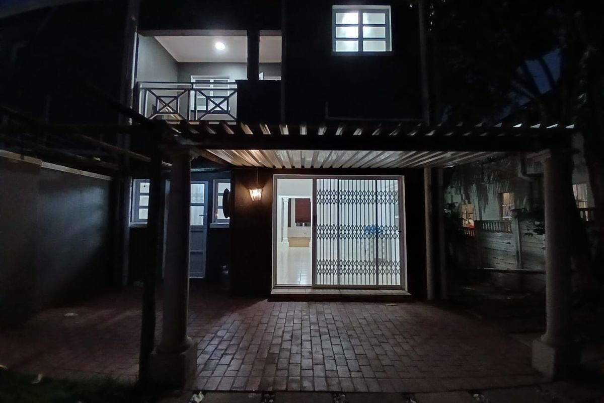 Rear exterior view of the property featuring a covered brick-paved patio, a security gate entrance, and an upper-level balcony at night.
