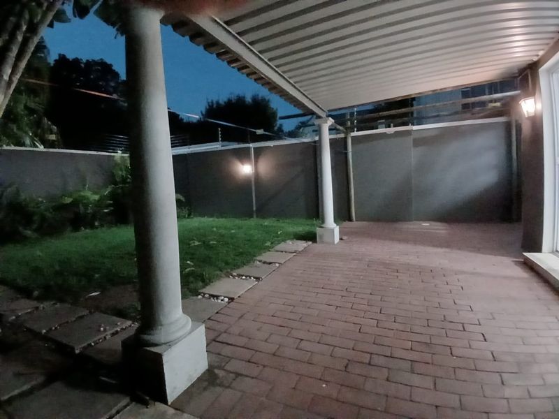 A covered patio area featuring brick paving, structural columns, and a view of the rear garden and boundary wall.