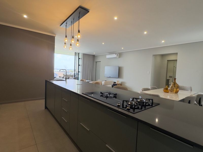Modern open-plan living area featuring a sleek kitchen island with a gas hob, recessed lighting, and neutral tiled flooring throughout.