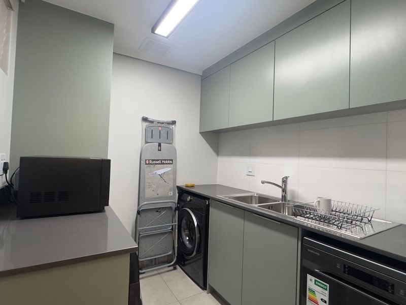 Modern laundry room featuring sage green cabinetry, stone countertops, stainless steel sink, and integrated washing machine with clean tiled finishes.