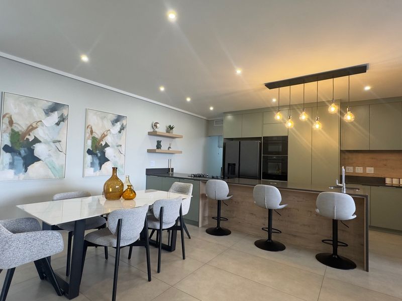 Modern open-plan kitchen and dining area featuring sleek cabinetry, integrated appliances, a functional kitchen island with breakfast bar, and contemporary pendant lighting.