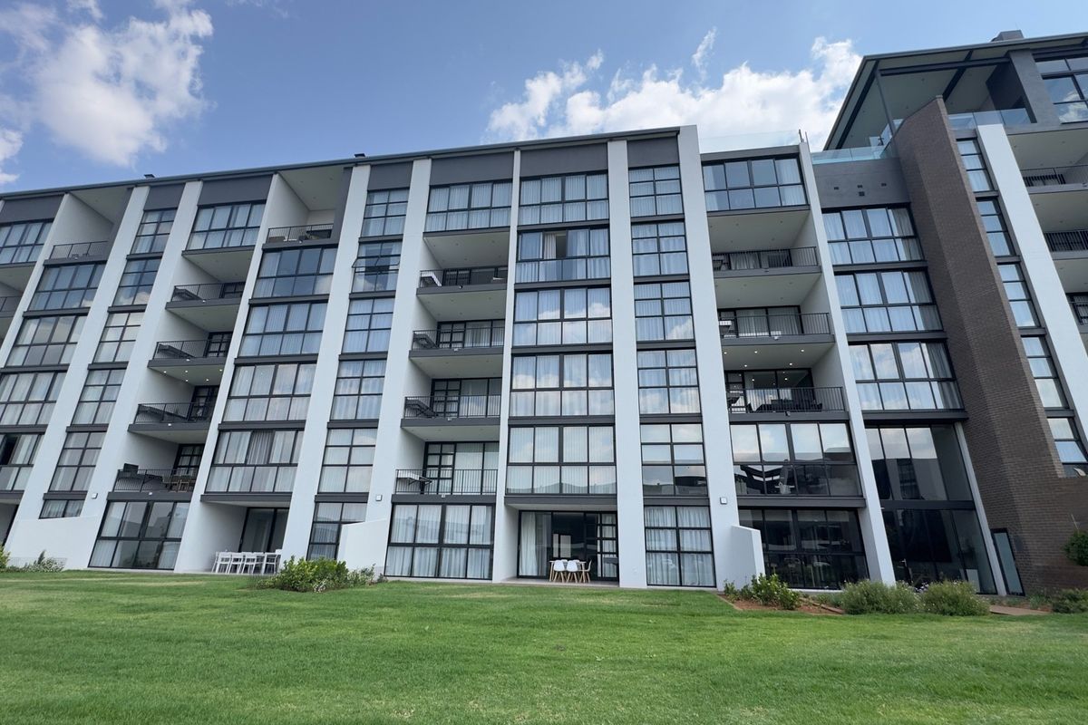 A modern multi-story apartment building exterior featuring extensive glass facades and private balconies overlooking a well-maintained lawn area.