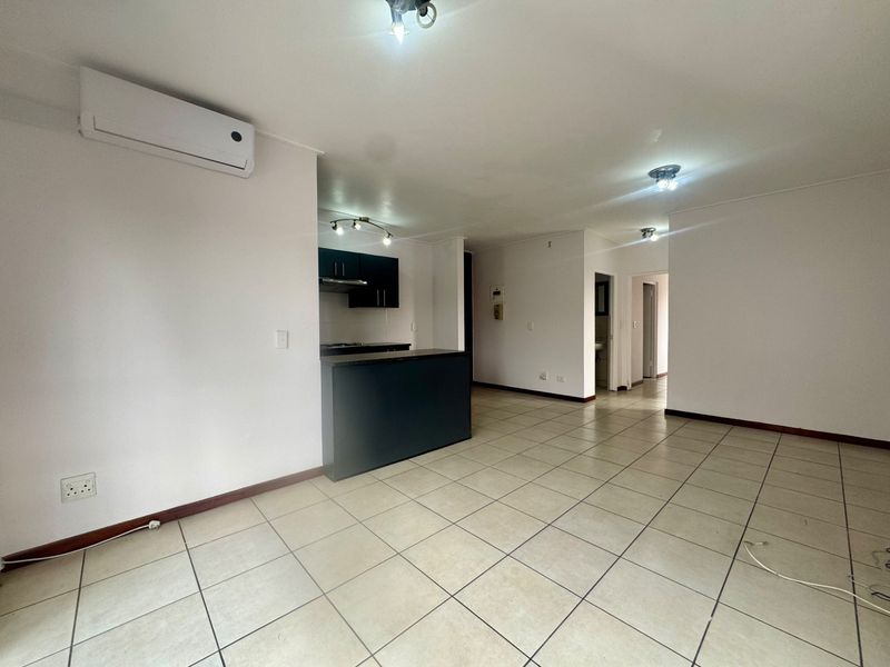 Spacious open-plan living area featuring tiled flooring, a wall-mounted air conditioning unit, and a compact kitchen with built-in cabinetry and breakfast bar.