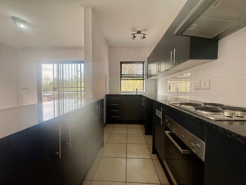 Modern kitchen featuring dark cabinetry, granite countertops, built-in oven, electric hob, and tiled flooring, with an open-plan layout leading to a balcony.
