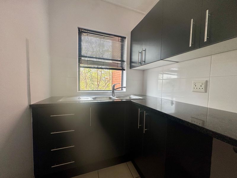 A compact, modern kitchen featuring dark cabinetry, granite countertops, a stainless steel double sink, and a tiled backsplash with natural light.