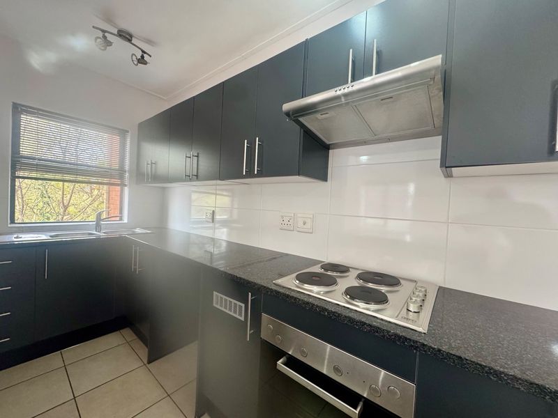 Modern kitchen featuring dark cabinetry, granite countertops, built-in oven, electric hob, extractor fan, and tiled flooring with ample natural light.