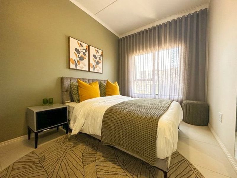 A well-maintained bedroom featuring neutral tiled flooring, soft green accent wall, and large window with floor-to-ceiling curtains providing natural light.