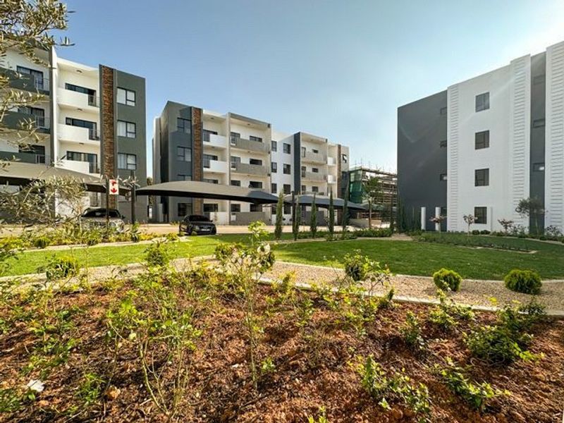 A well-maintained apartment complex courtyard featuring landscaped gardens, paved pathways, and covered parking bays between modern multi-story residential buildings.