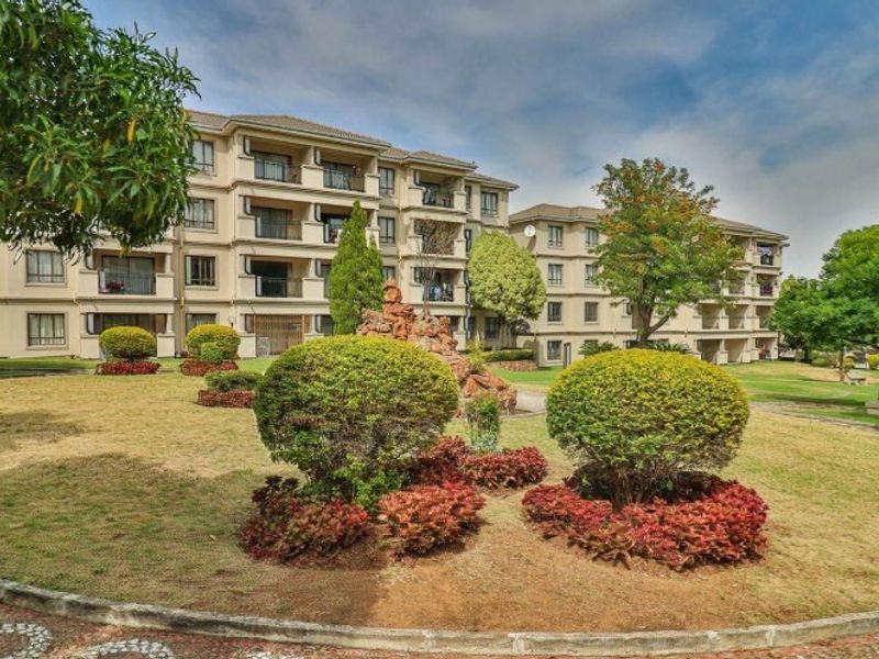 A view of the apartment complex exterior featuring a well-maintained landscaped garden, paved walkways, and multiple residential balconies.
