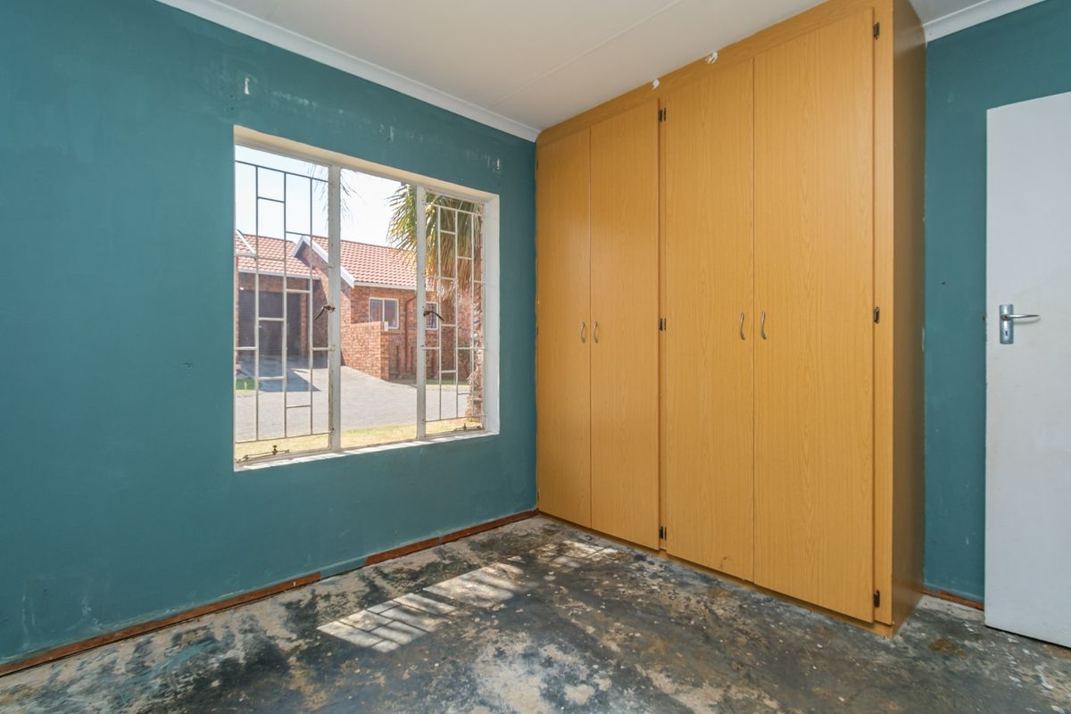 An empty bedroom featuring built-in wooden cupboards, security bars on the window, and flooring in need of replacement or refurbishment.