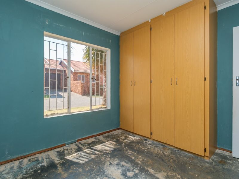 An empty bedroom featuring built-in wooden cupboards, security bars on the window, and flooring in need of replacement or refurbishment.