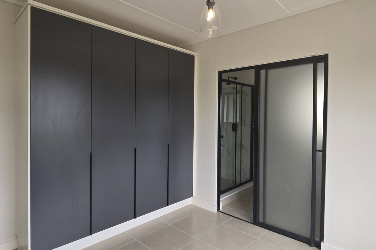 A modern bedroom featuring sleek dark built-in cupboards, neutral tiled flooring, and a contemporary frosted glass sliding door leading to an en-suite bathroom.