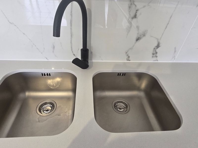 A modern kitchen sink area featuring a double stainless steel basin, matte black gooseneck tap, and elegant marble-look splashback with stone countertops.