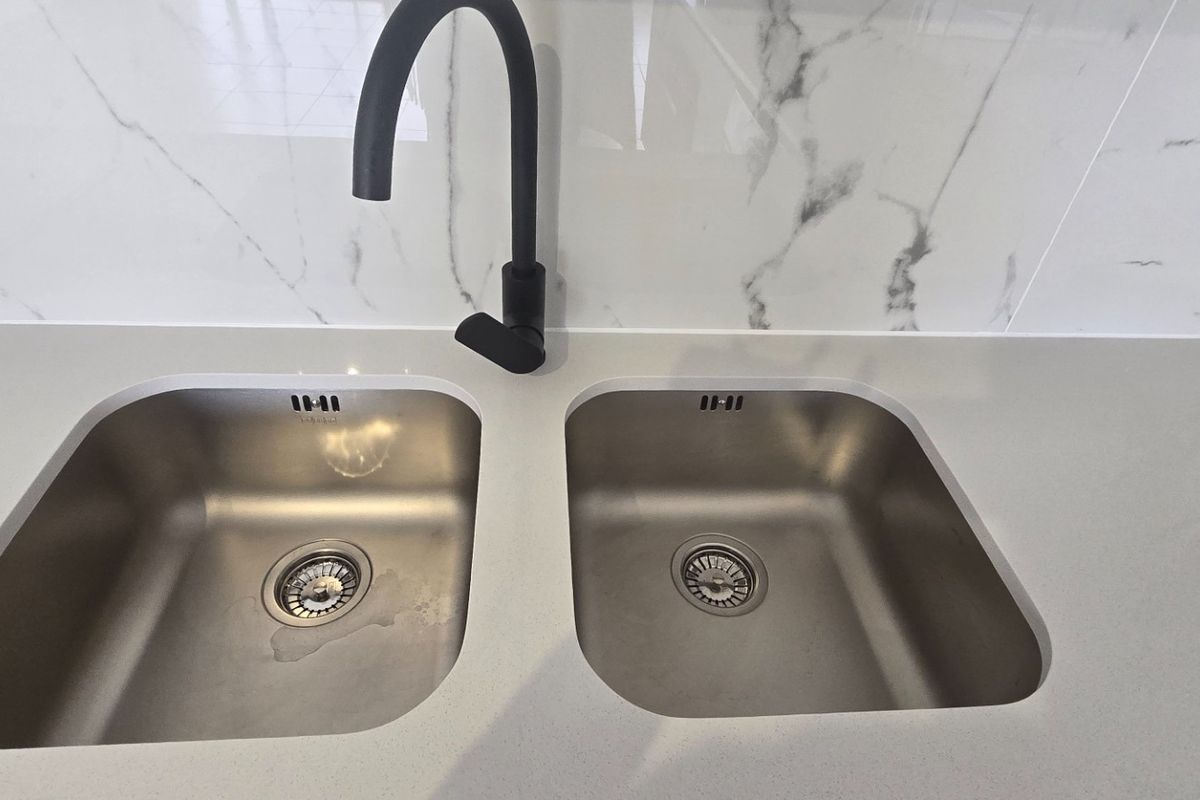 A modern kitchen sink area featuring a double stainless steel basin, matte black gooseneck tap, and elegant marble-look splashback with stone countertops.