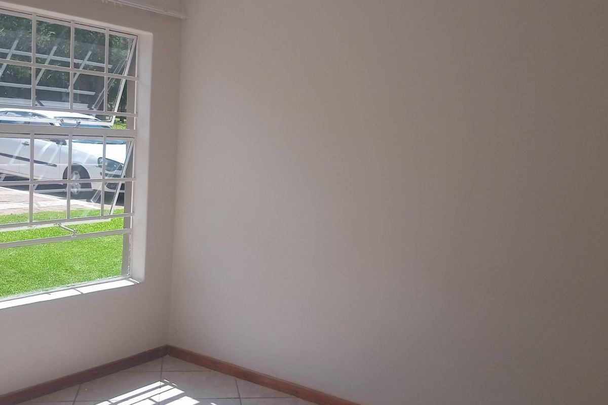 An empty living area featuring tiled flooring, neutral walls, and a large window with security bars overlooking the exterior grounds.