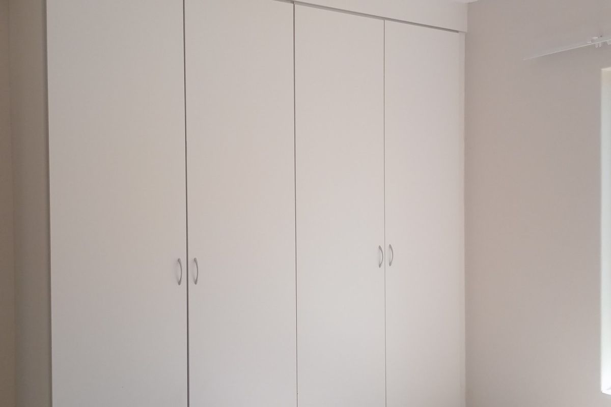 A clean, empty bedroom featuring neutral walls, built-in cupboards, and a window overlooking the exterior area.